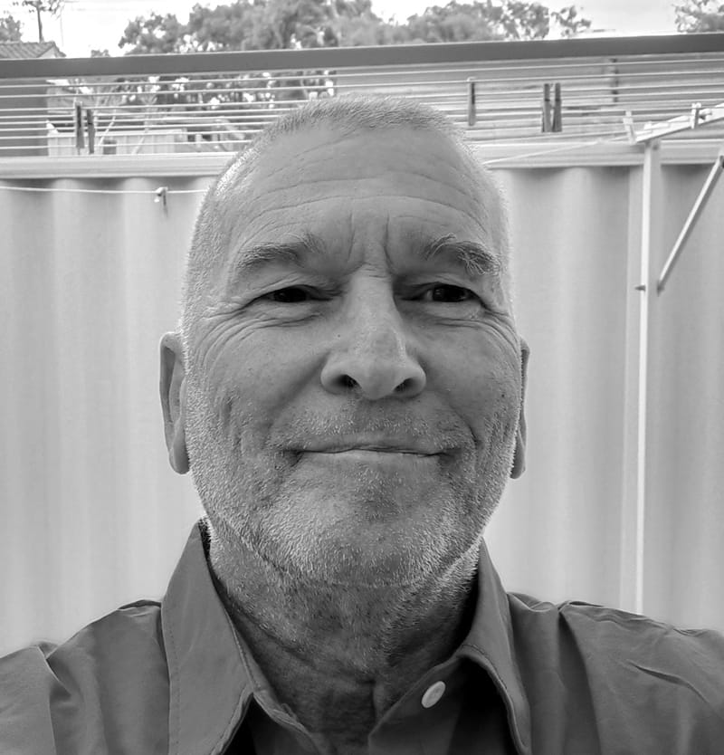 Man in collared shirt framed by fibrous fence and trees—black and white portrait of stillness and latent tension.